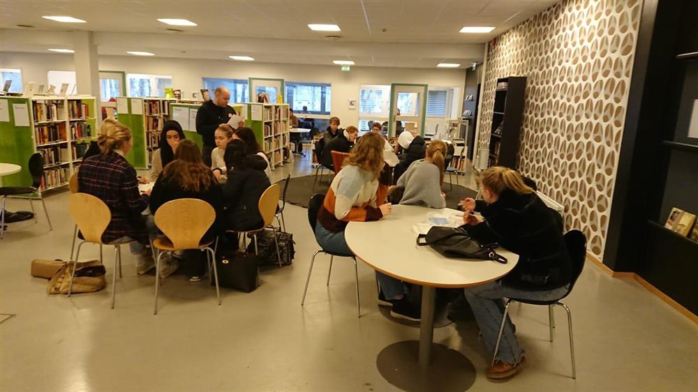 Miljøarbeider Sjur arrangerer quiz i midttimen. Elever sitter ved runde bord i biblioteket og løser quiz - Klikk for stort bilde