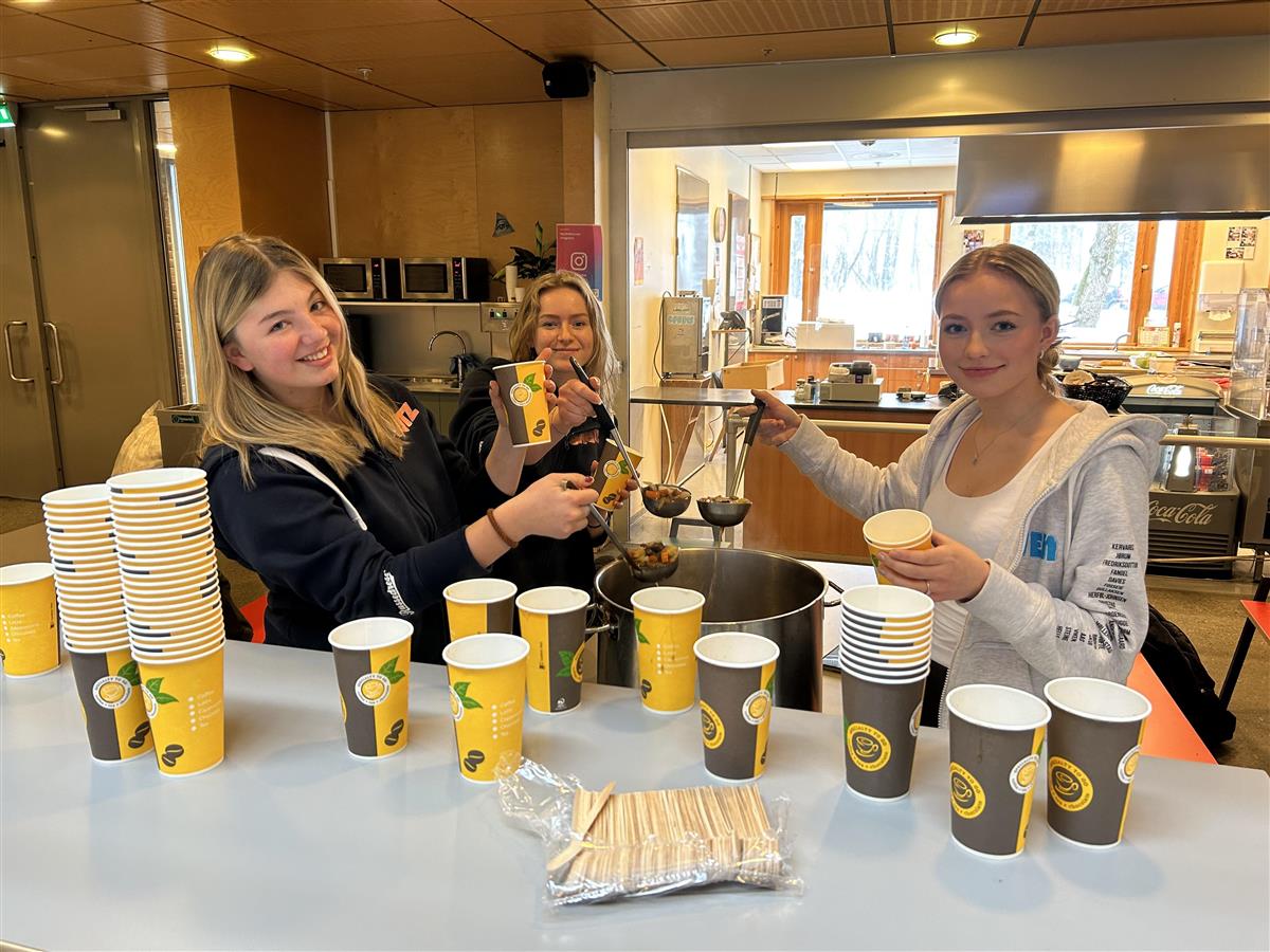 Tre blide jenter fra sosiale dager komitéen står ved en kjele og serverer den samiske retten Bidos til alle i kantina - Klikk for stort bilde