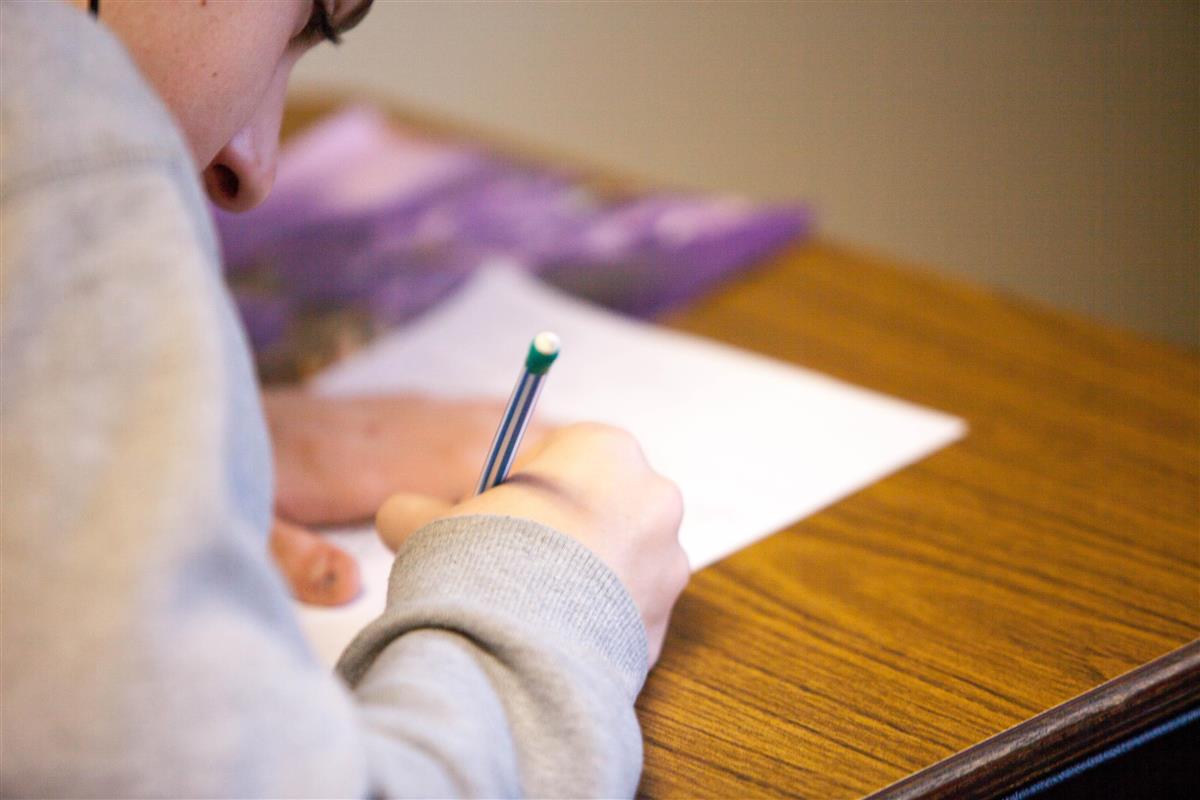 Elev med grå genser ses i skrå vinkel bakfra. Skriver med blyant på hvitt ark. - Klikk for stort bilde