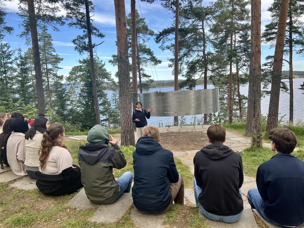 En gruppe ungdommer sitter og ser på minnesmerket over de drepte 22.07.11. på Utøya, mens en guide står foran minnesmerket og forteller. - Klikk for stort bilde