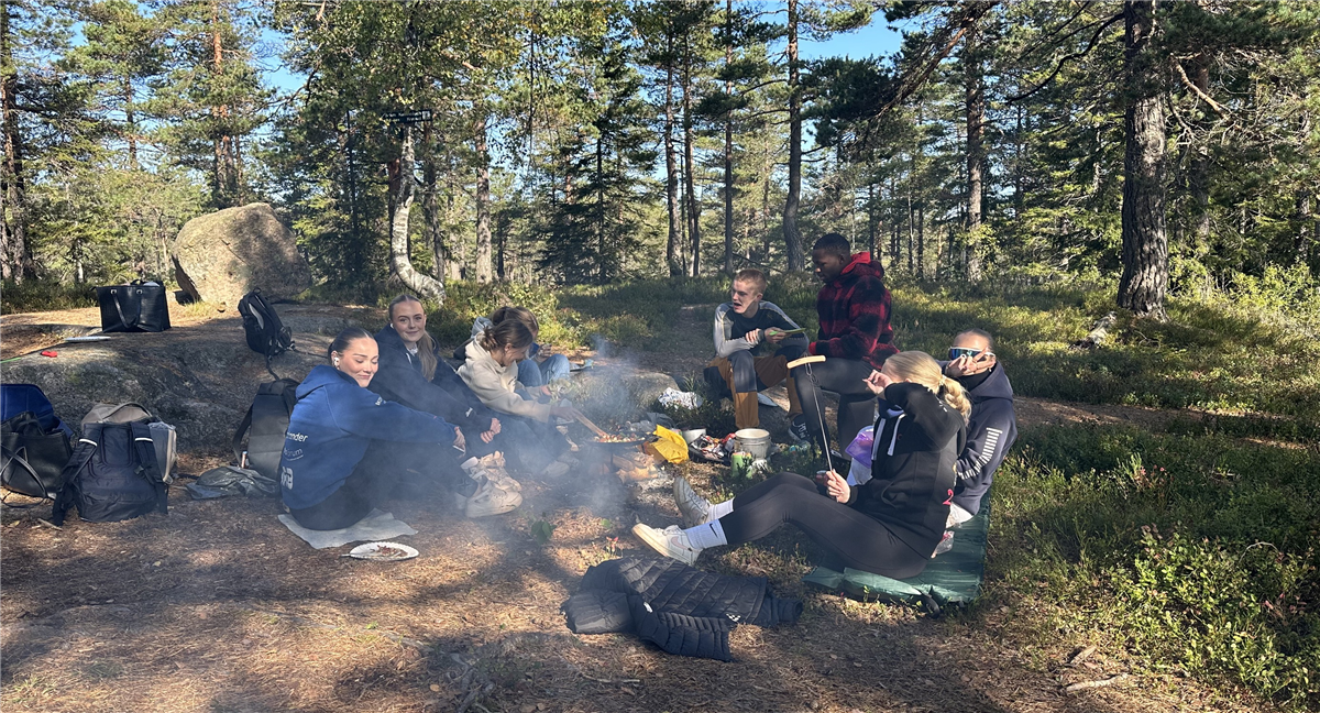 En gruppe ungdommer sitter rundt et bål i skogen og lager mat - Klikk for stort bilde