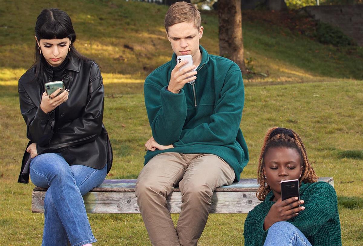Tre ungdommer sitter på hver sin mobil, jente og gutt på benk, jente på bakken. - Klikk for stort bilde