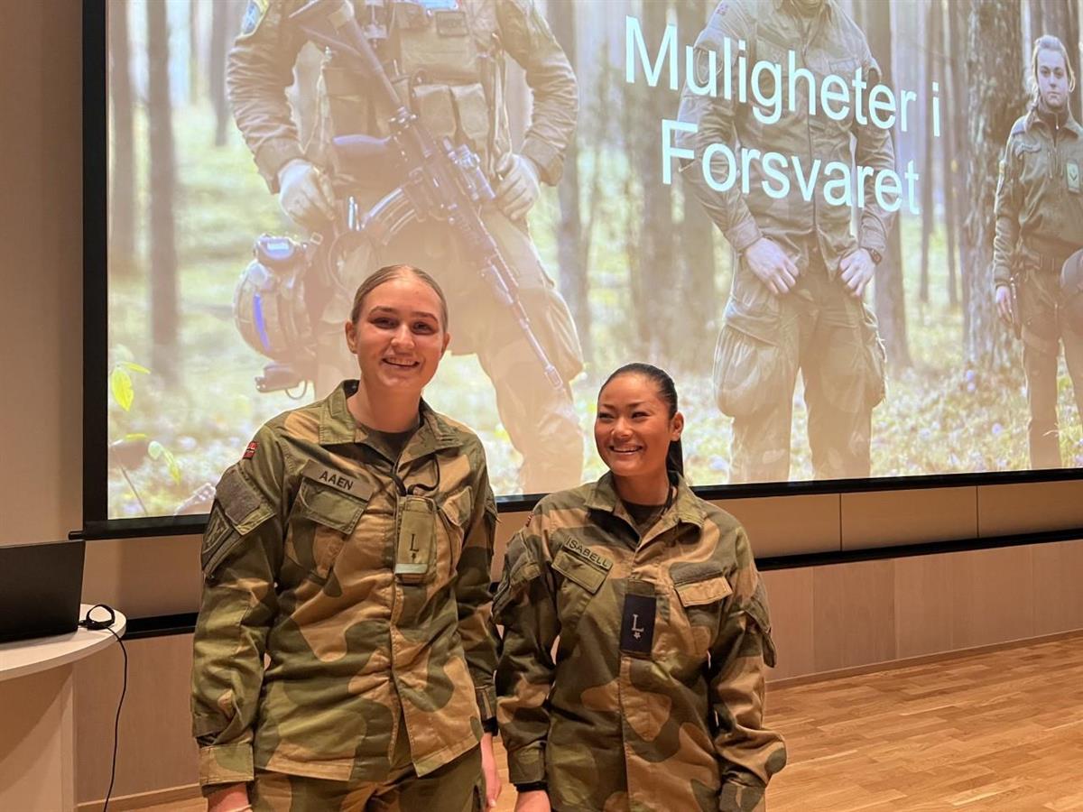 To unge kvinner i uniform står foran et bilde hvor det står 'Muligheter i Forsvaret'. - Klikk for stort bilde