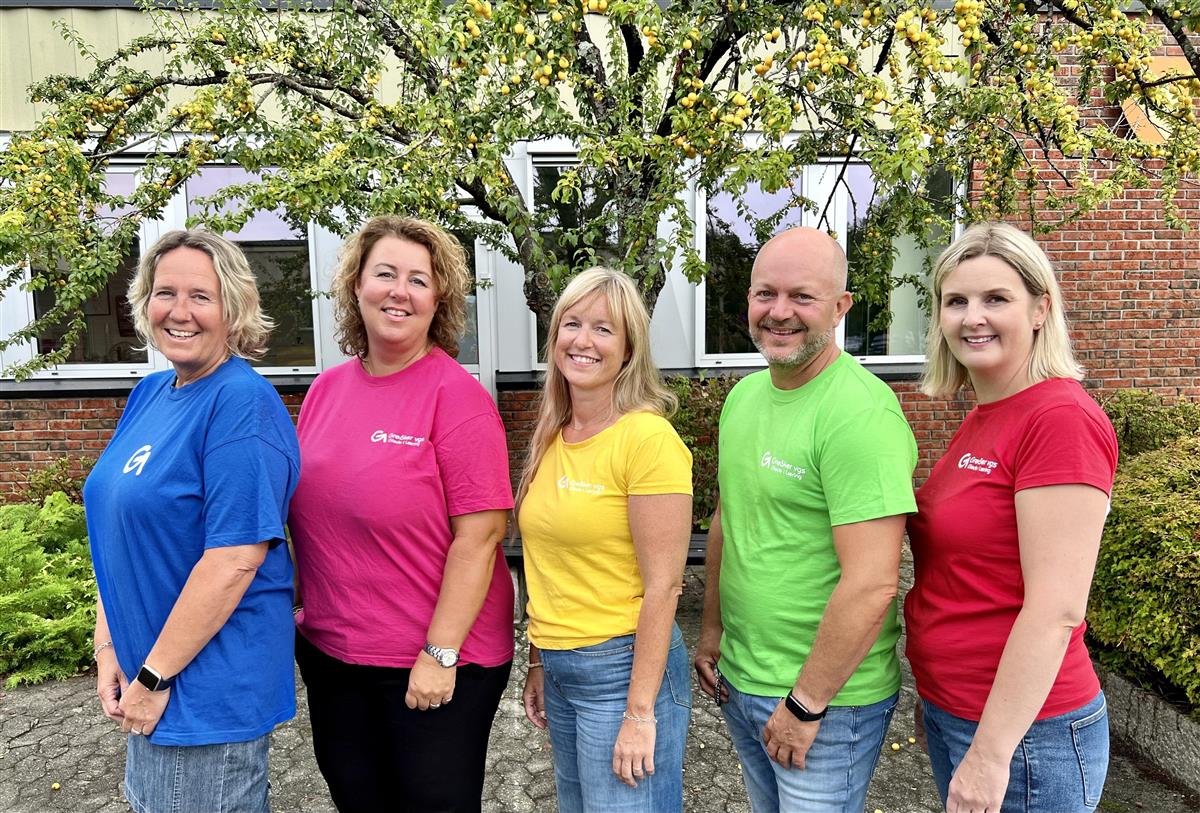 Fire kvinner og en mann i fargesterke t-skjorter fotografert foran et skolebygg og et tre.  - Klikk for stort bilde