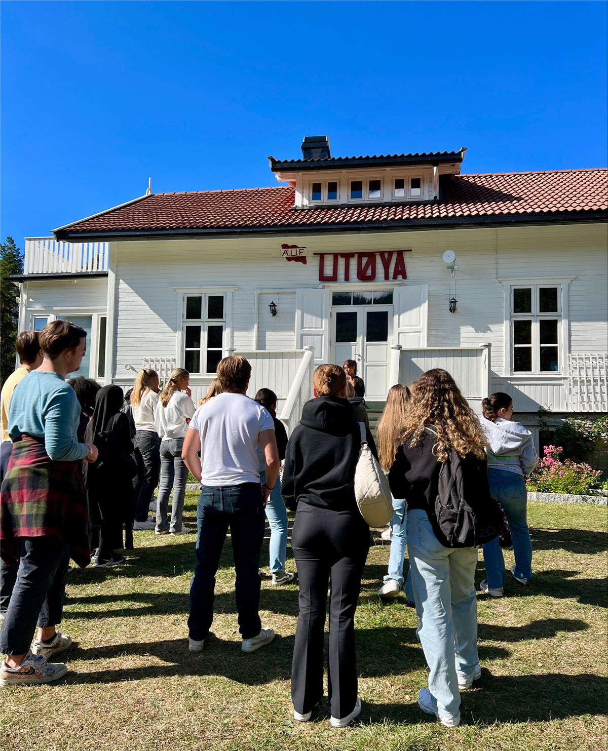 Det er blå himmel og en gruppe elever står med ryggen til fotografen på en gressplen og hører på en dame som snakker.  De står foran et hus med skriften "AUF Utøya" over inngangsdøren. - Klikk for stort bilde