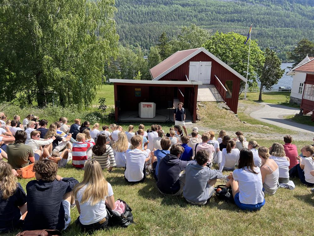 En gruppe ungdommer sitter i en gressbakke og hører på en som forteller fra en talerstol.  - Klikk for stort bilde